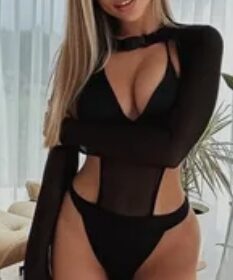 Woman in a black sheer bodysuit with cutouts posing in a bright room by sheer curtains, looking at the camera with a soft smile.